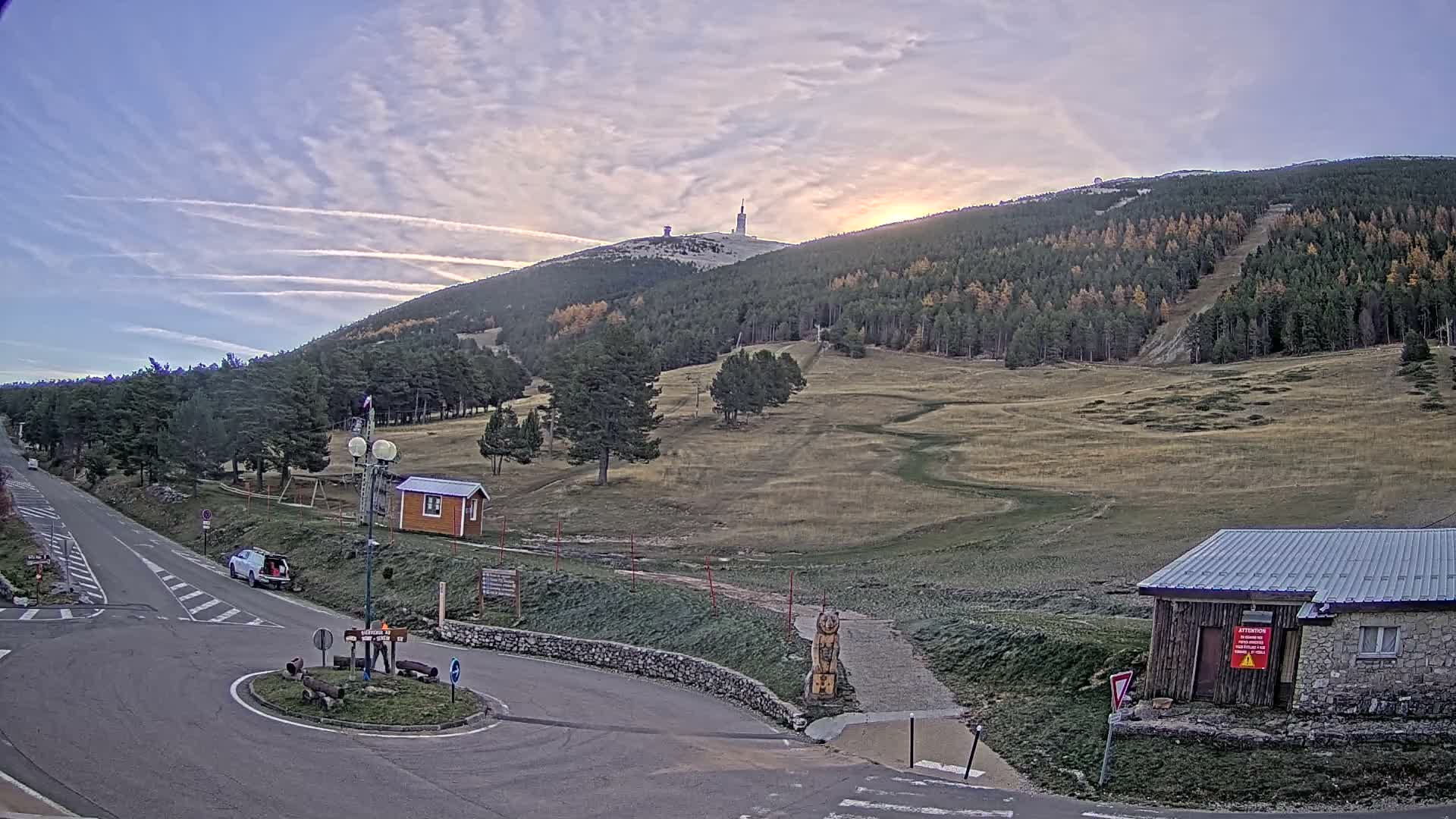 Under a partly cloudy sky with a low sun, a winding road with a roundabout and a white van passes through a mountainous autumn landscape featuring dense pine forests, grassy ski slopes, and various buildings, with a communications tower visible on the distant peak.