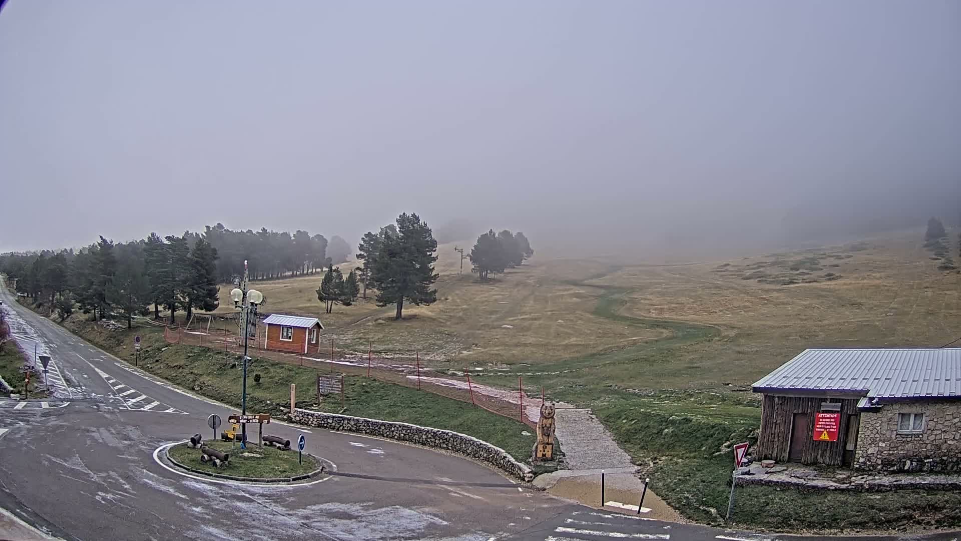 A misty, overcast mountain landscape is seen, with a wet asphalt road and roundabout in the foreground, flanked by grassy fields, scattered pine trees, and rustic buildings disappearing into a thick fog.