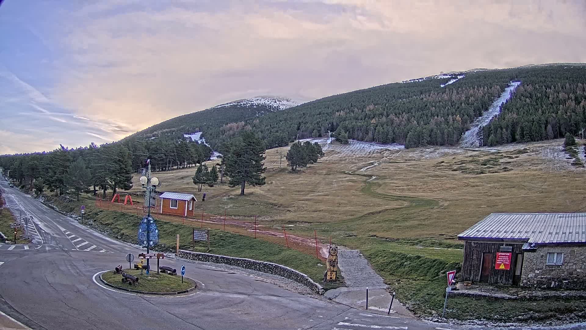 A cold, overcast day reveals a mountainous landscape featuring a road leading to a roundabout, dense evergreen forests, grassy slopes with patches of snow on higher elevations and along trails, and scattered small buildings.