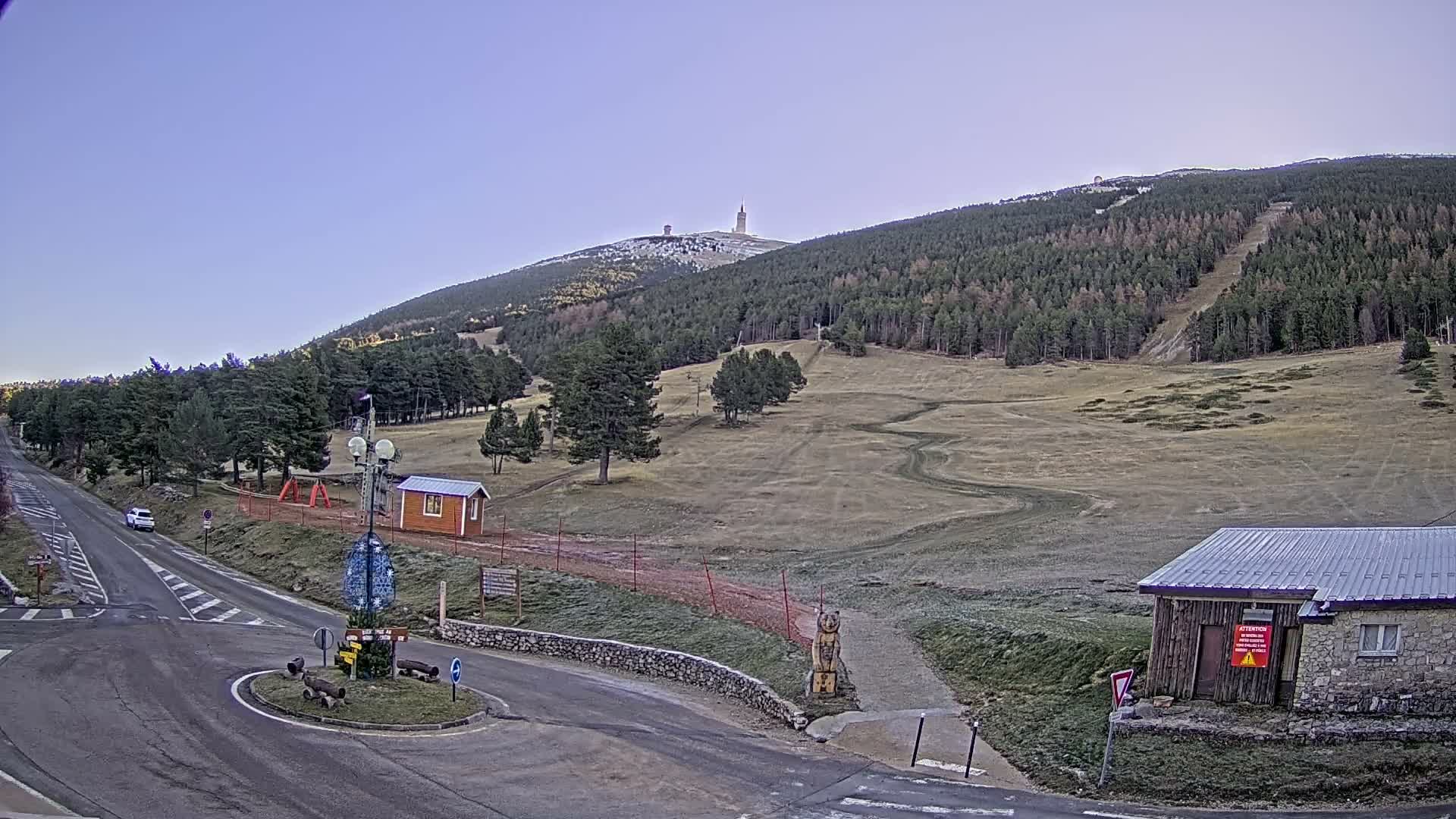 A cold, overcast day reveals a mountainous landscape featuring a road leading to a roundabout, dense evergreen forests, grassy slopes with patches of snow on higher elevations and along trails, and scattered small buildings.