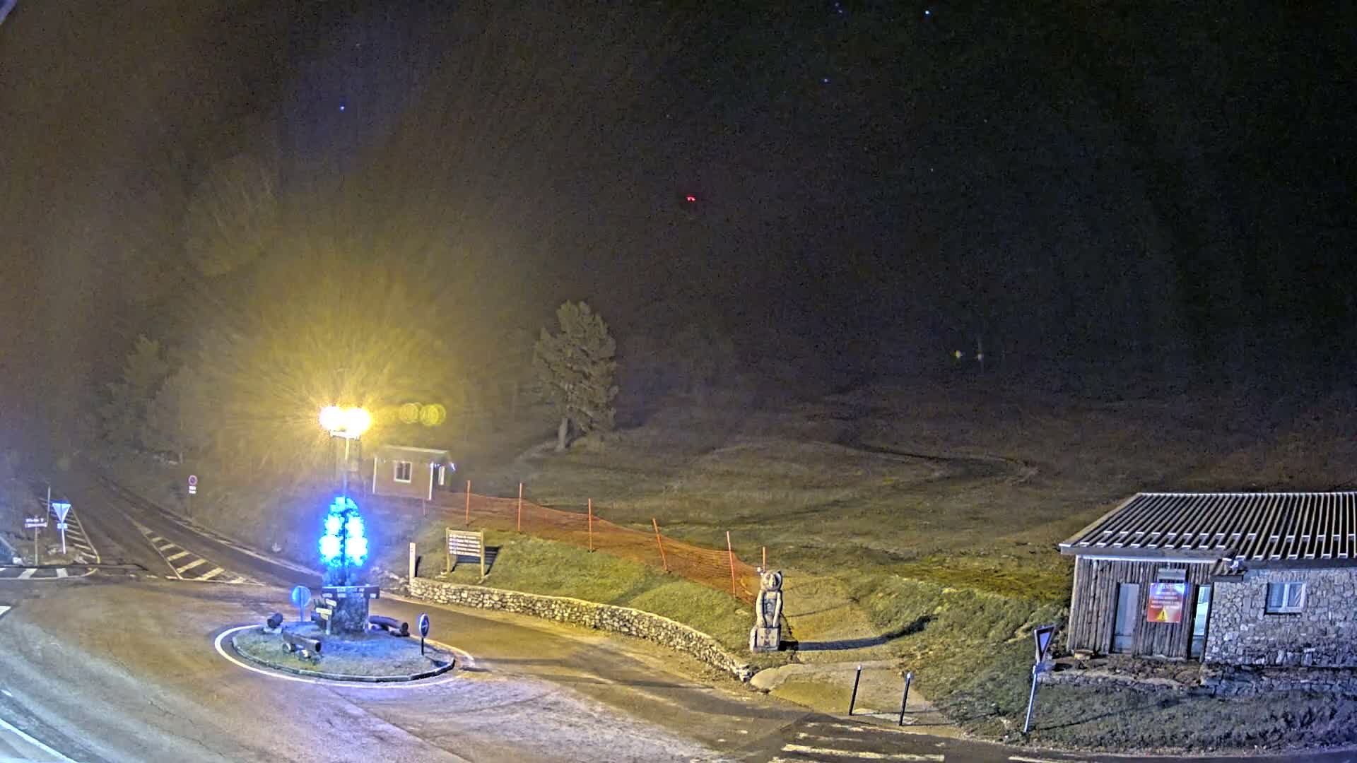 A cold, overcast day reveals a mountainous landscape featuring a road leading to a roundabout, dense evergreen forests, grassy slopes with patches of snow on higher elevations and along trails, and scattered small buildings.