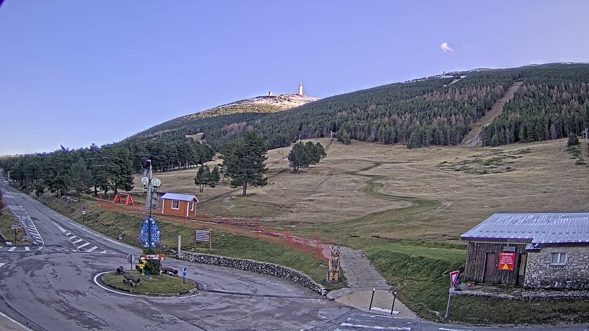 A cold, overcast day reveals a mountainous landscape featuring a road leading to a roundabout, dense evergreen forests, grassy slopes with patches of snow on higher elevations and along trails, and scattered small buildings.