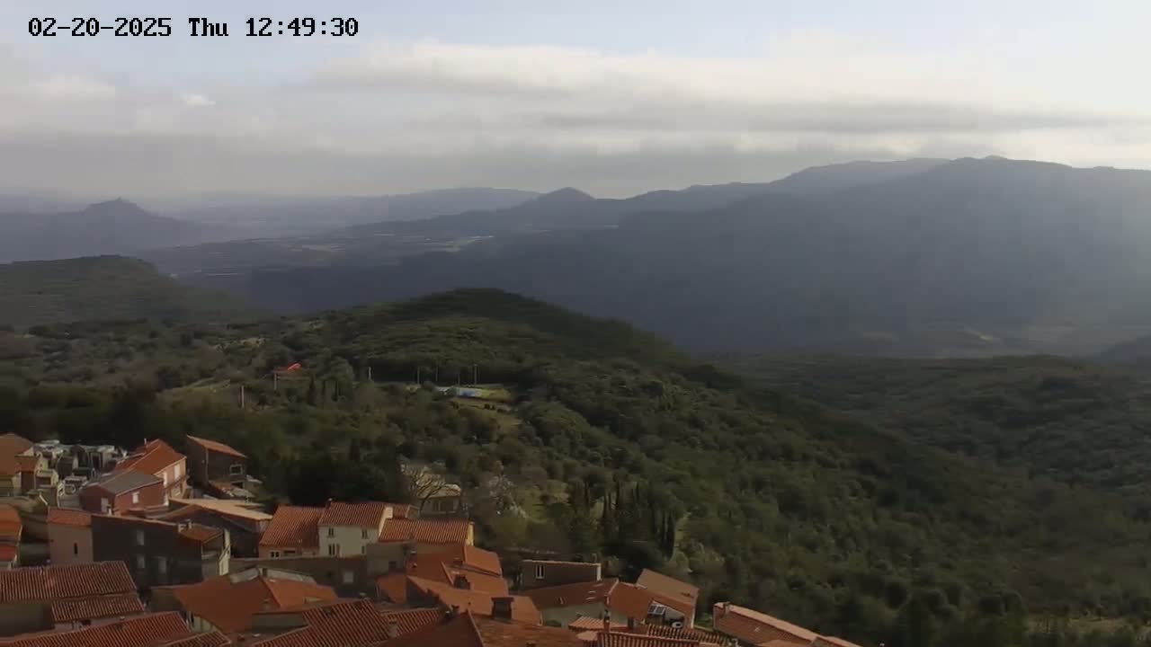 A village with red-tiled roofs sits atop a hill overlooking a vast, hazy mountain range under a mostly cloudy sky.
