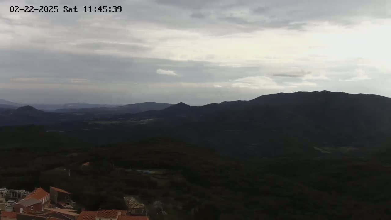 A village nestled in a dark green, mountainous landscape under a mostly cloudy sky.
