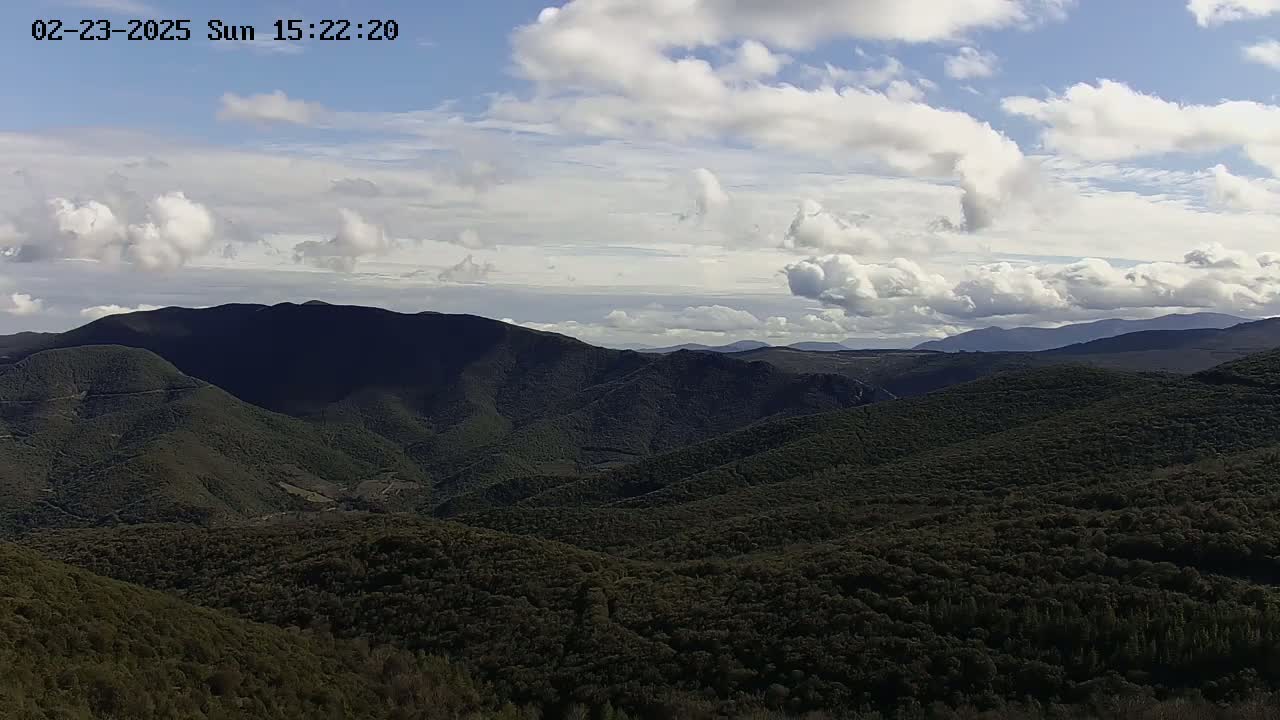 A partly cloudy sky overlooks a vast expanse of dark green, rolling hills and mountains.