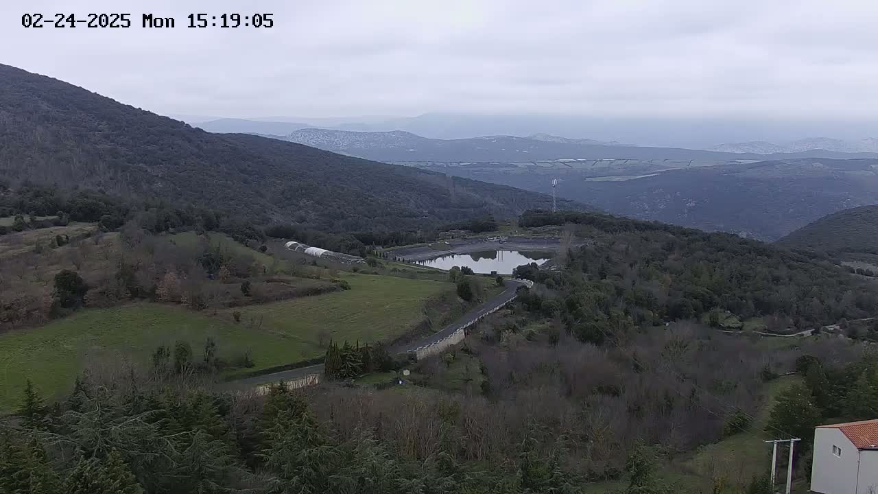 A winding road snakes through a hilly landscape of green fields and dark forests under a cloudy sky, culminating in a small reservoir reflecting the overcast light.