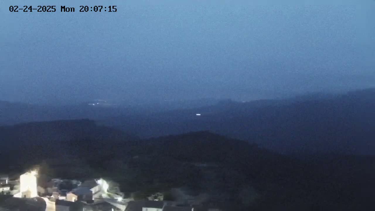 A nighttime view of a small village nestled in dark hills under a dusky blue sky.