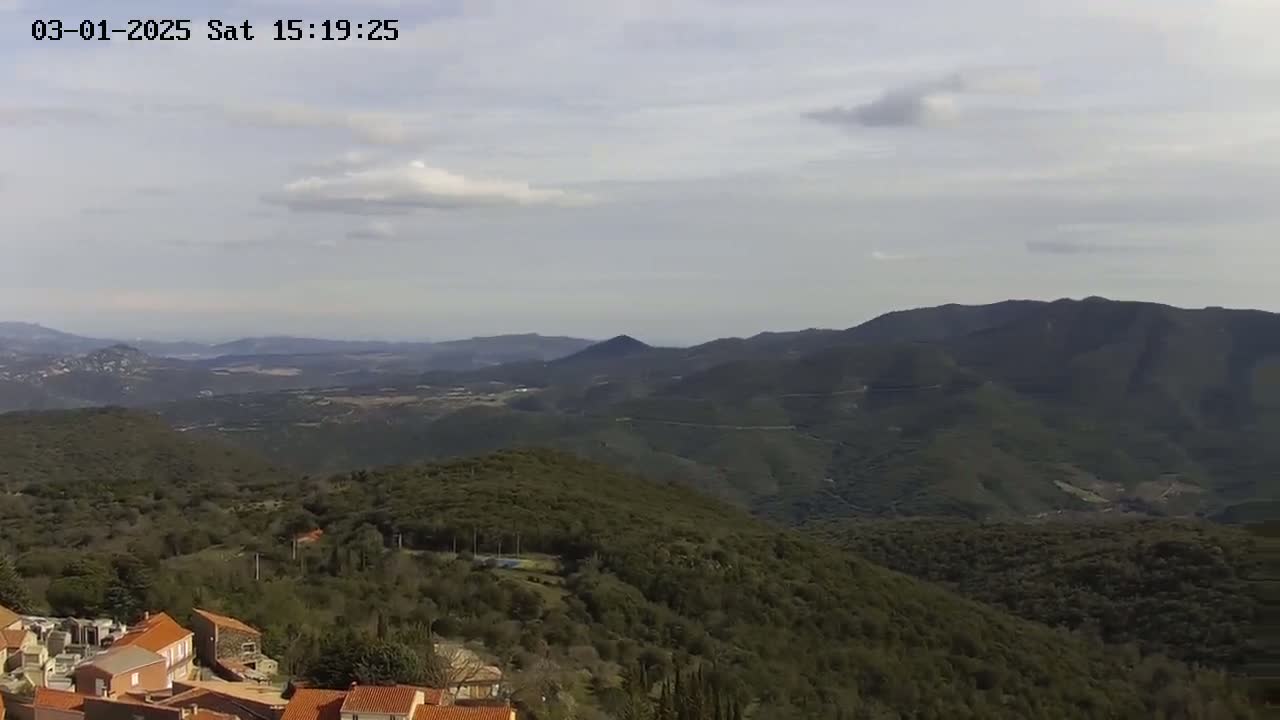 A partly cloudy day reveals a view of a small village nestled in green, mountainous terrain, overlooking a vast expanse of rolling hills and distant mountains.