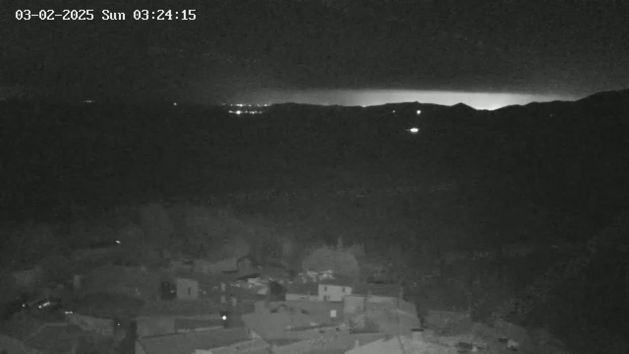 A nighttime, low-light view shows a small village nestled below a distant horizon line illuminated by faint lights and a possible lightning strike.