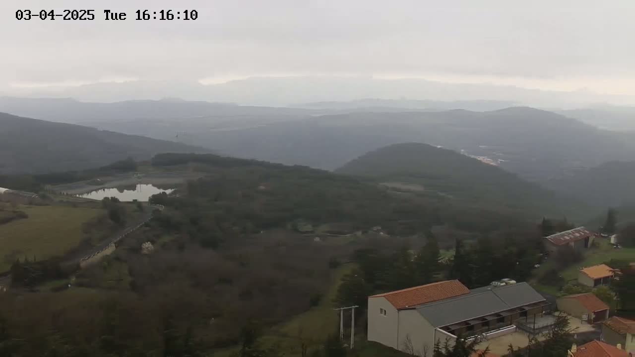 A hazy view overlooking a hilly landscape with a small body of water, a few buildings, and sparsely wooded areas under a gray sky.