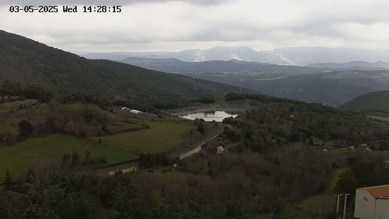 A hilly landscape under an overcast sky shows a small reservoir nestled among dark green, sparsely populated hills, with snow-capped mountains in the distance.