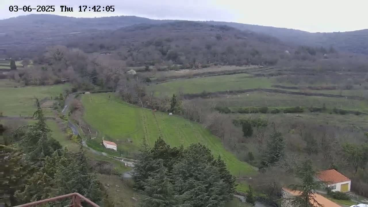 A high-angle view of a rural landscape on an overcast day shows rolling green hills, sparsely wooded areas, and a few small buildings.