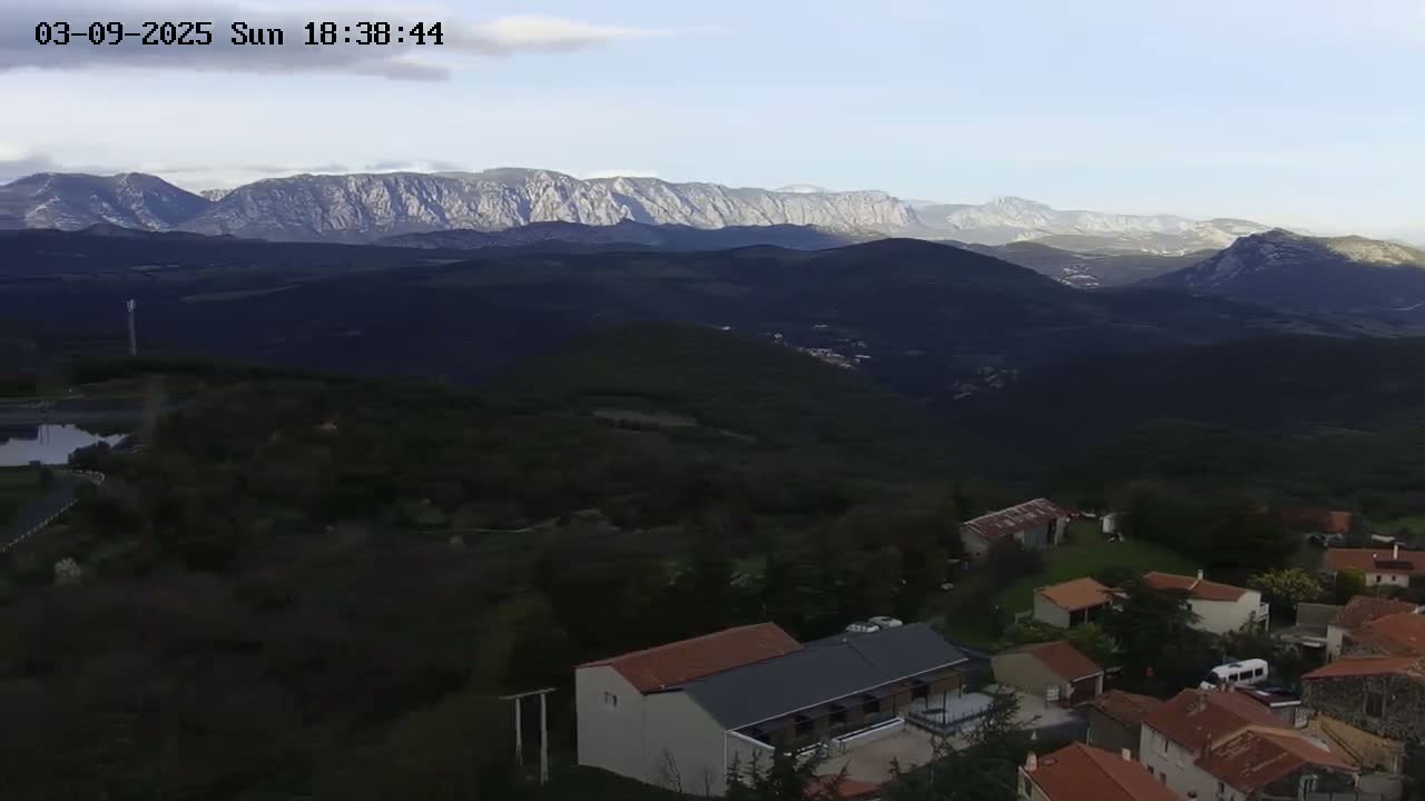 A small village nestled in a valley, with a mountain range in the background under a partly cloudy sky.
