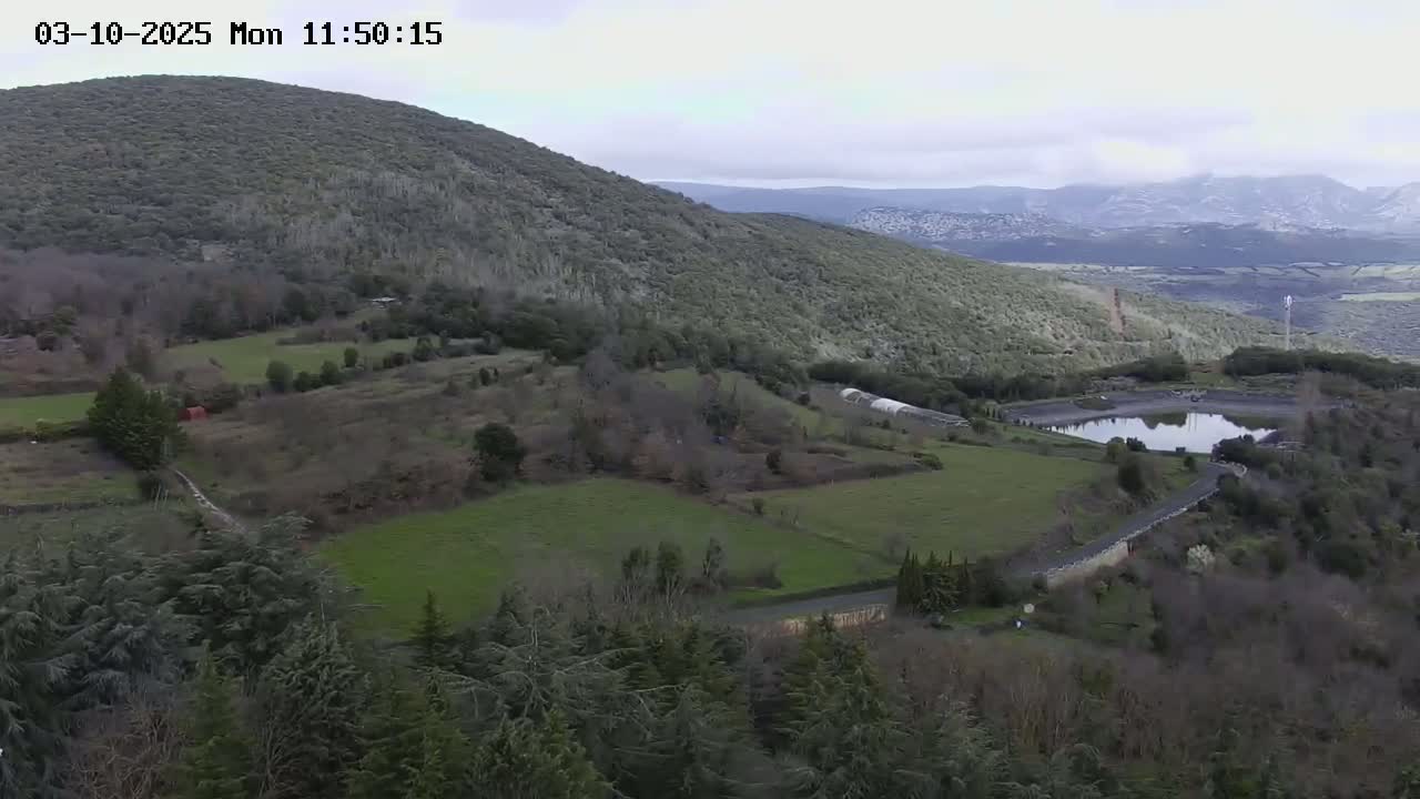 A high-angle view shows a hilly landscape under an overcast sky, with fields, trees, and a small body of water.