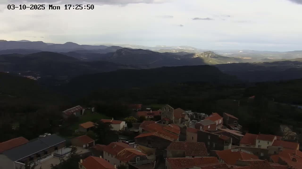 A small village with red-tiled roofs nestled in a mountain valley under a partly cloudy sky.