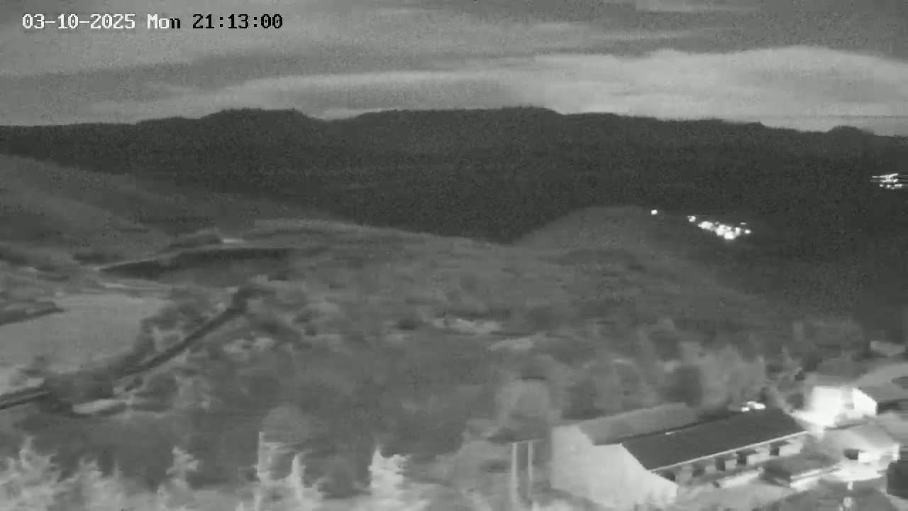 A nighttime, low-light view shows a hilly landscape with a few buildings and some lights in the distance under a partly cloudy sky.