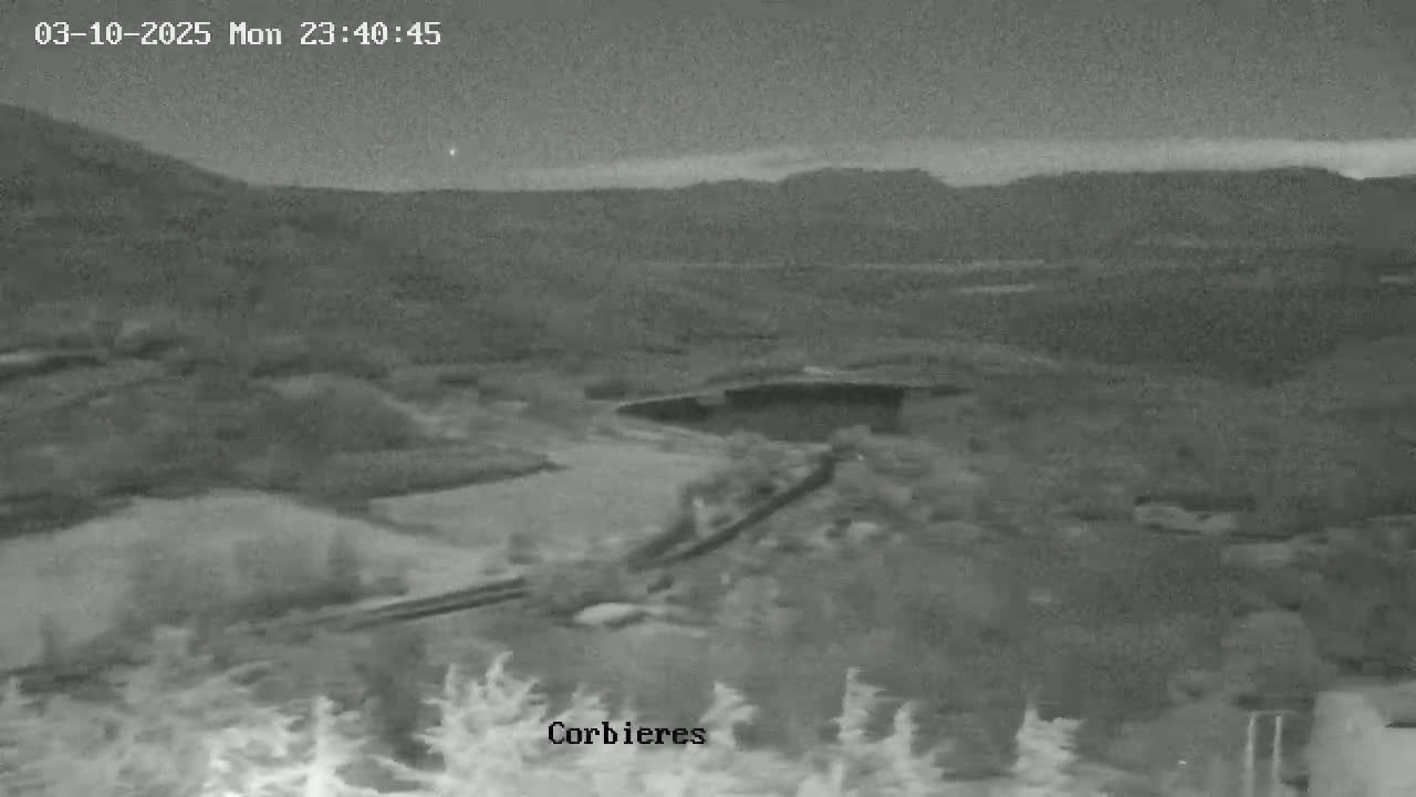 A nighttime, low-light image shows a hilly landscape with a bridge visible in the middle ground under a mostly clear sky.