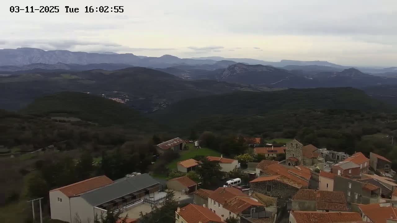 An aerial view shows a small village nestled in a hilly landscape under an overcast sky, with mountains visible in the distance.