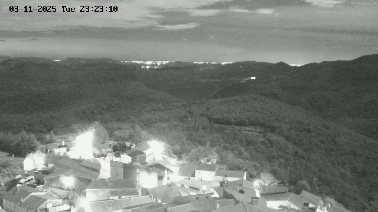 A nighttime, grayscale view shows a small village nestled in a valley, with distant city lights visible on the horizon under a partly cloudy sky.