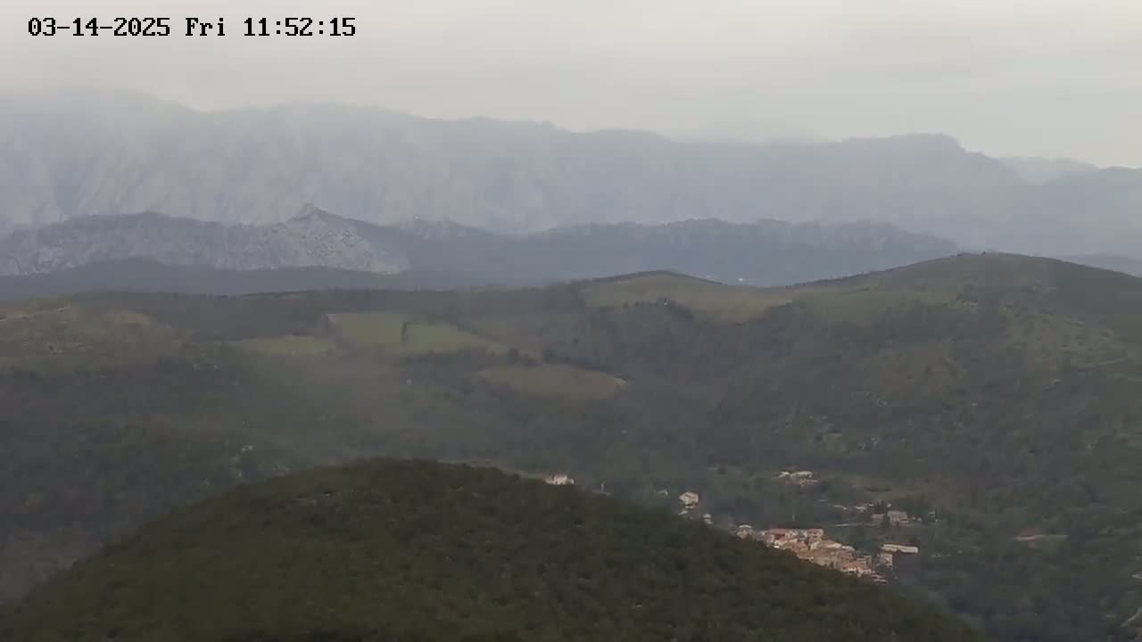 A hazy, overcast view shows rolling hills and distant mountains, with a small village nestled in the valley below.