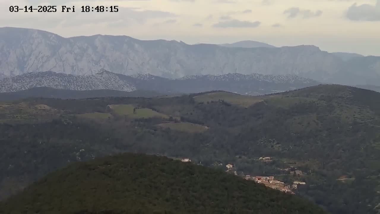 A long view of rolling hills and mountains under an overcast sky shows a small village nestled among the lower hills.