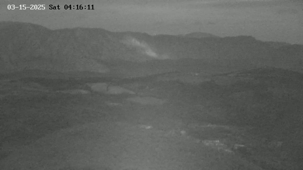 A hazy, low-light view of a mountainous landscape shows a possible plume of dust or smoke rising from a mountainside.