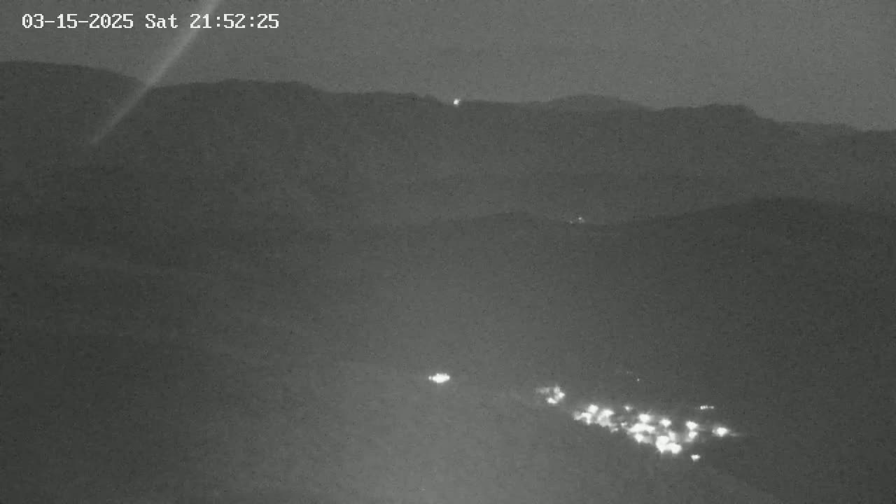 A nighttime long shot shows a valley with a few scattered lights below dark mountains under a clear, dark sky.