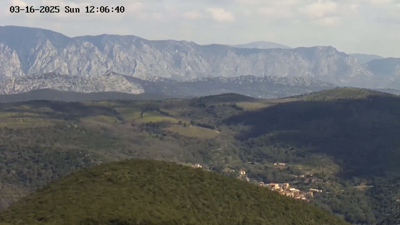 A long-range view shows a mountainous region under a mostly clear sky, with a small village nestled in the foothills below a range of light gray peaks.