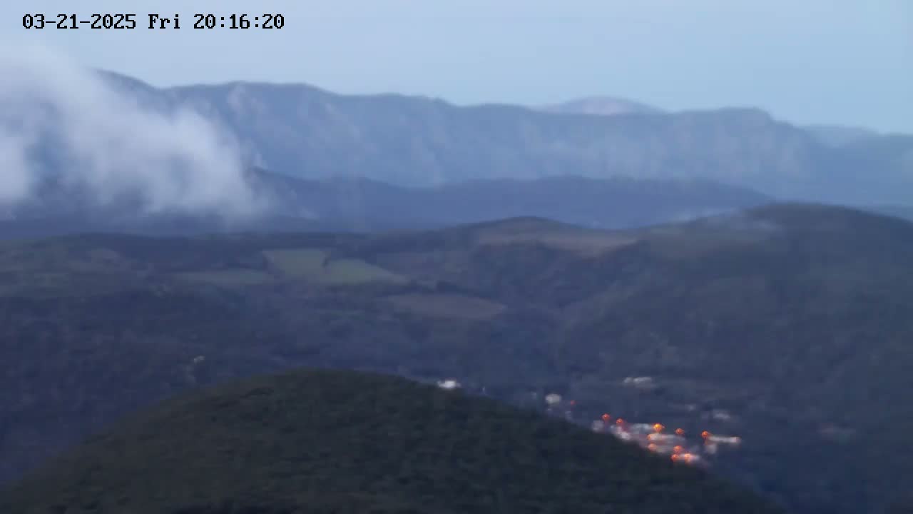 A hazy, mountainous landscape at dusk shows a small cluster of orange lights in a valley below a large cloud of white mist clinging to a distant mountain range.