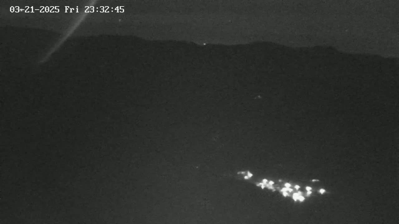 A nighttime, low-light view shows a cluster of lights in a valley below a dark mountain range under a clear sky.