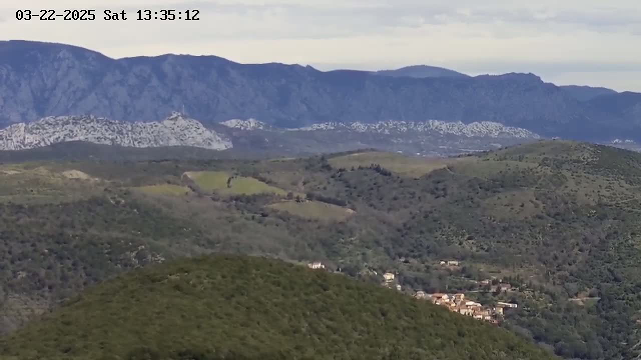 A distant view of a mountainous landscape under a cloudy sky, showing a small village nestled among rolling green hills in the foreground.
