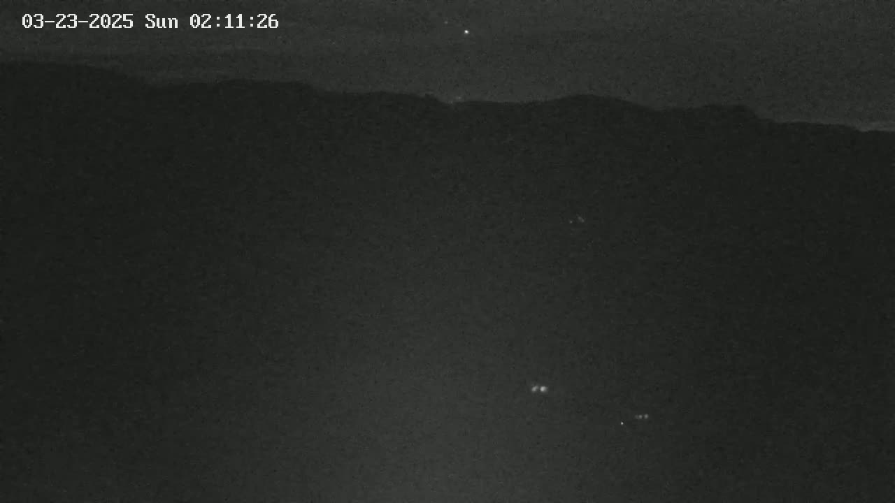 A nighttime, low-light image shows a dark, mountainous landscape with a few faint lights visible in the lower portion of the frame.