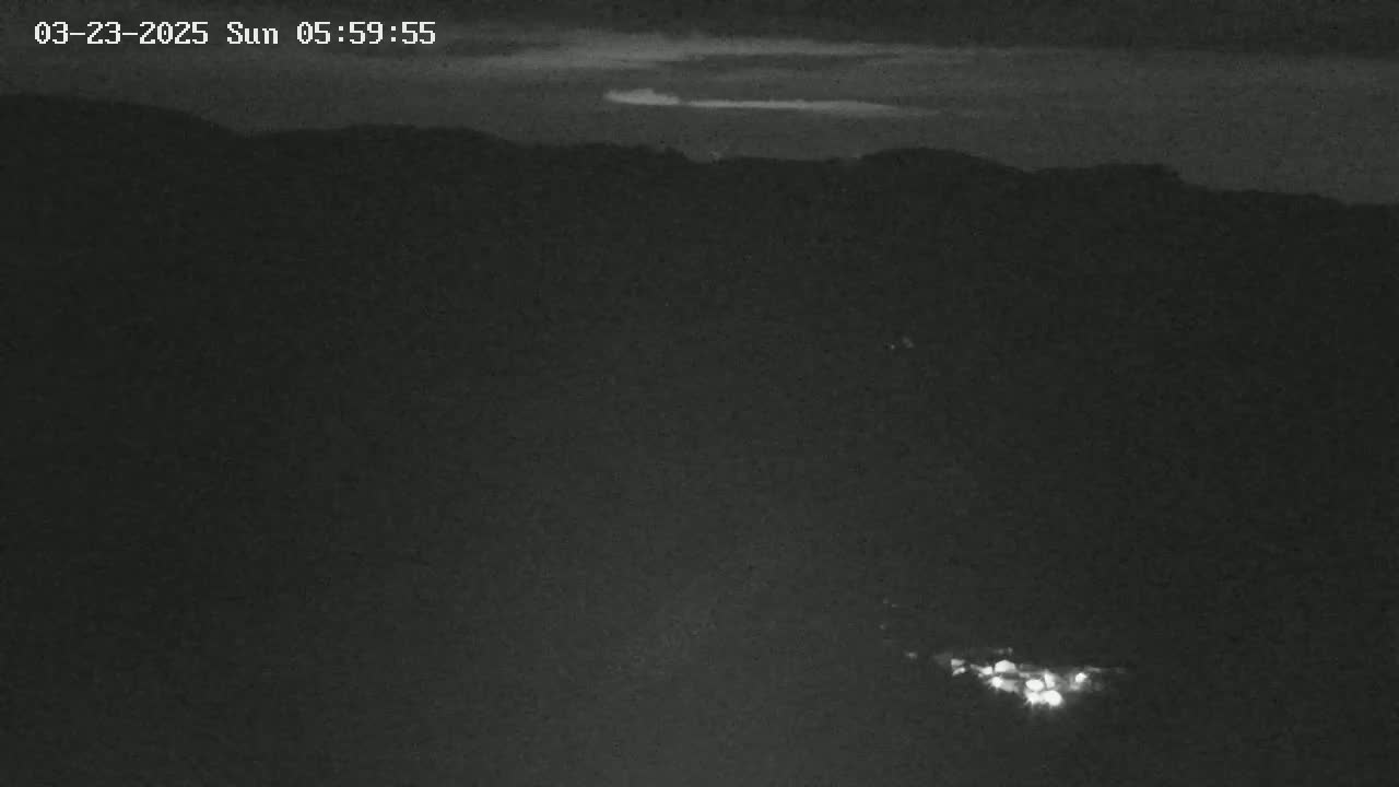 A low-light nighttime image shows a dark landscape with faint lights in the lower portion and a slightly brighter sky suggesting the very beginning of dawn.