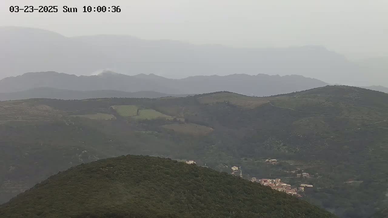 A hazy view of rolling green hills and a small village nestled amongst them under an overcast sky.
