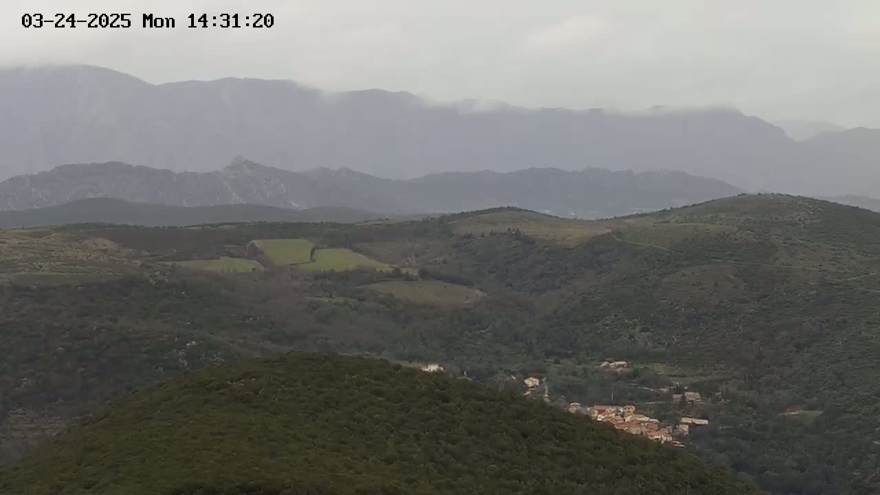 A hazy, overcast day reveals a rolling landscape of green hills and a small village nestled in a valley, with distant mountains shrouded in mist.