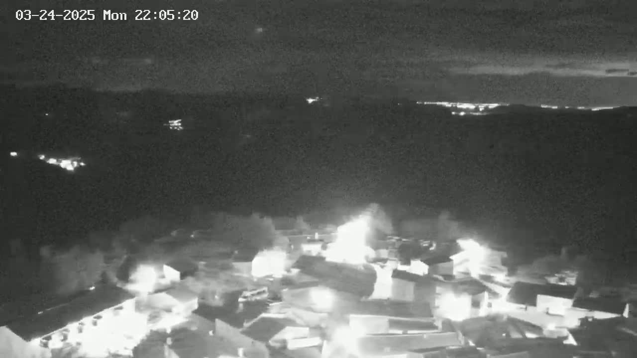 An aerial nighttime view shows a town with multiple bright lights and what appears to be a fire or intense light sources amongst the buildings under a dark, mostly clear sky.