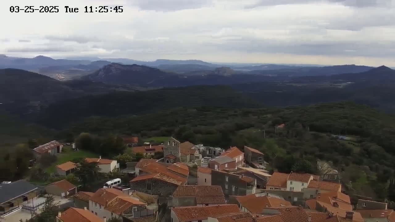 An aerial view shows a small village nestled in a mountainous region under a cloudy sky.