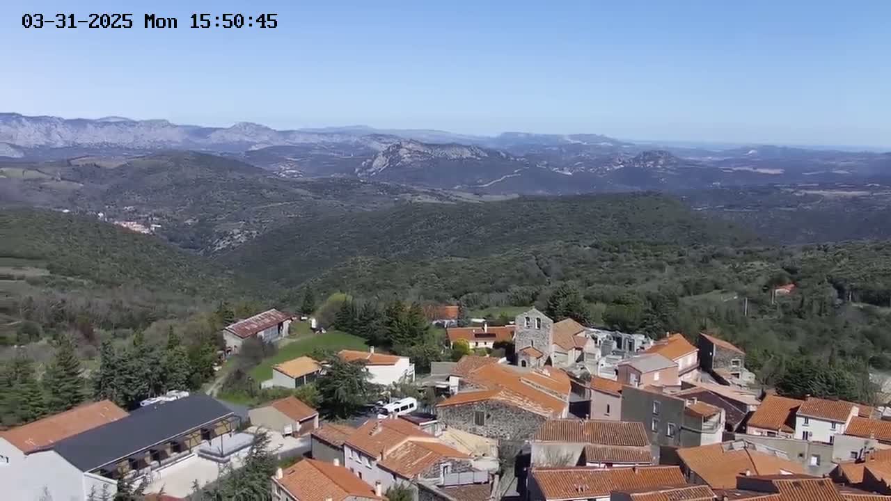 An aerial view shows a small village of terracotta-roofed buildings nestled in a verdant hillside, overlooking a vast mountain range under a clear blue sky.