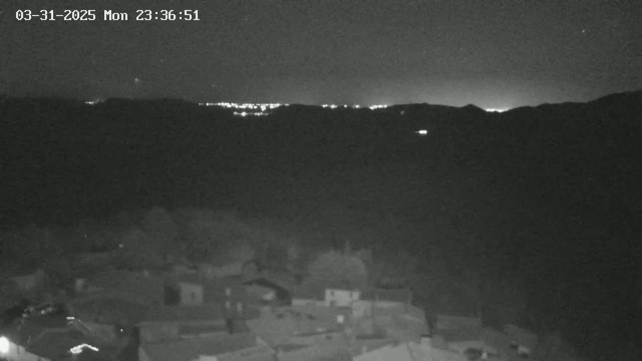 A nighttime view shows a village nestled in a valley, with distant city lights illuminating a mountain range under a clear, dark sky.