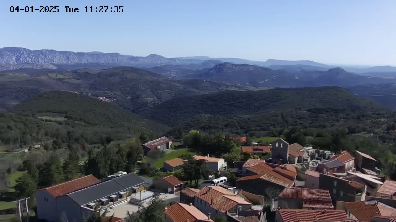 An aerial view shows a small village nestled in a valley, surrounded by rolling hills and mountains under a clear blue sky.