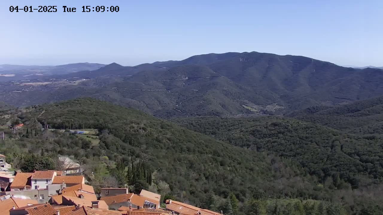 A village with orange-tiled roofs sits on a hillside overlooking a vast expanse of green, rolling hills and mountains under a clear blue sky.