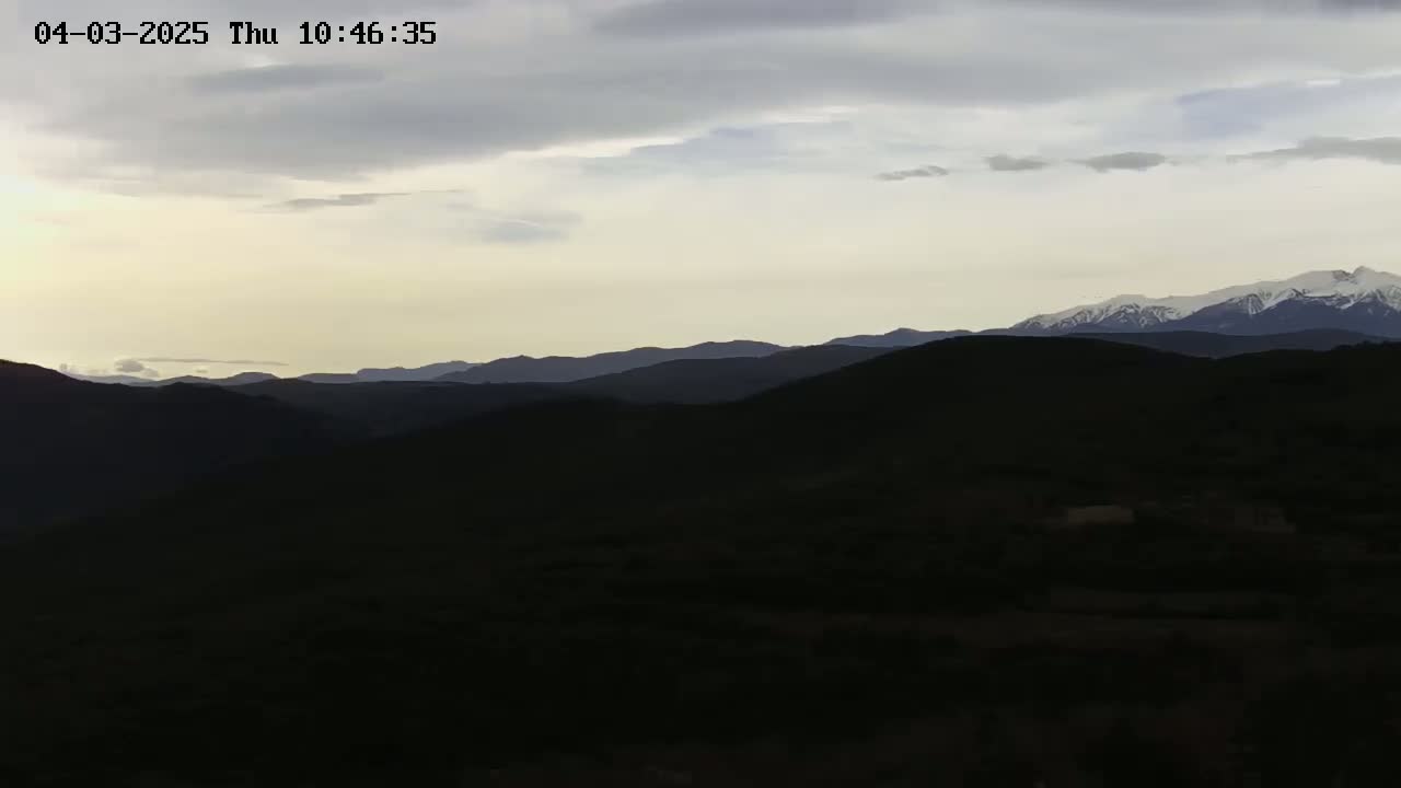 A mostly cloudy sky overlooks a dark, mountainous landscape with snow-capped peaks in the distance.