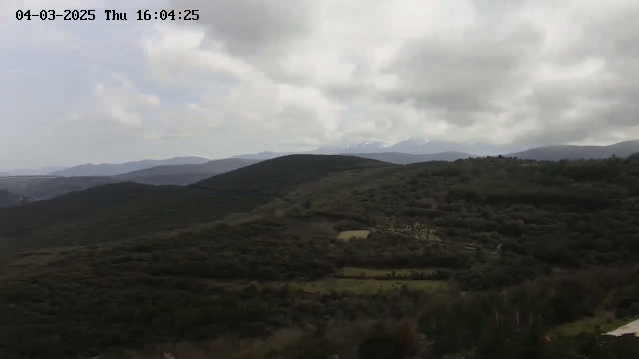 A view of a mountainous landscape under a cloudy sky shows dark green rolling hills and distant snow-capped mountains.