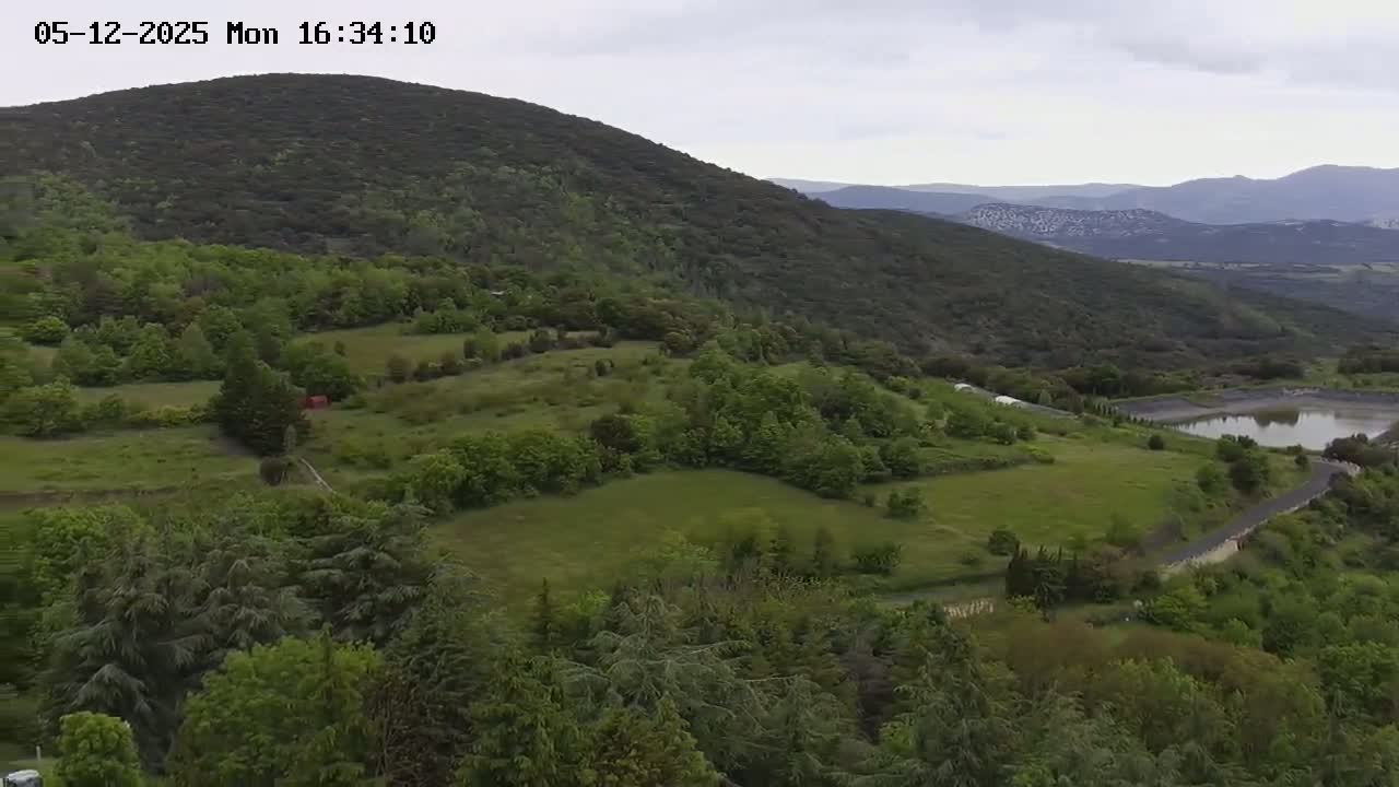 An aerial view shows a verdant landscape of rolling hills, lush green forests, and a small body of water under an overcast sky.