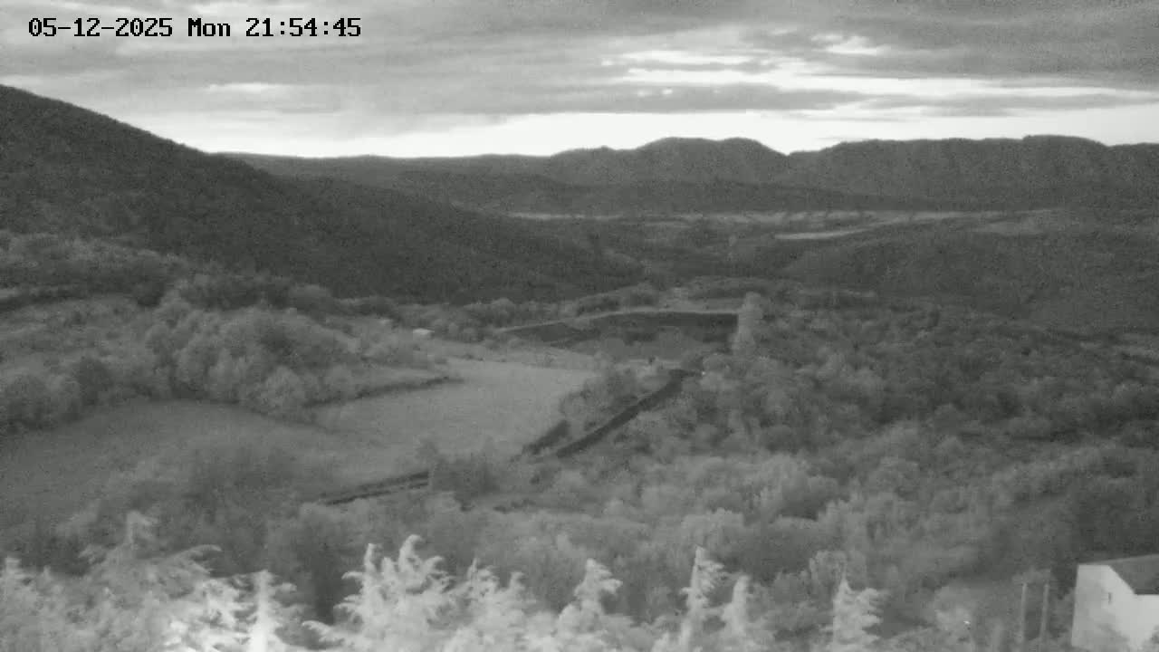 A high-angle grayscale view shows a hilly landscape under a cloudy twilight sky, featuring fields, forests, and a winding road.