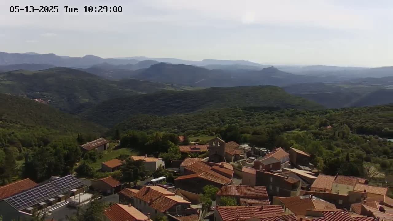 An aerial view shows a village of red-roofed buildings nestled in a green, mountainous region under a mostly clear sky.