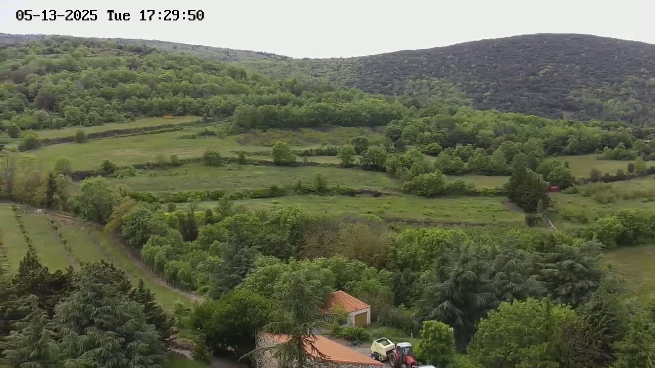 An aerial view shows a lush green landscape of fields and forests under an overcast sky, with a small building and some farm equipment visible near the bottom.
