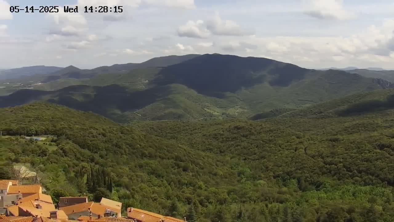 A partly cloudy day reveals a vista of rolling green hills and mountains, with a small village of orange-roofed buildings in the foreground.