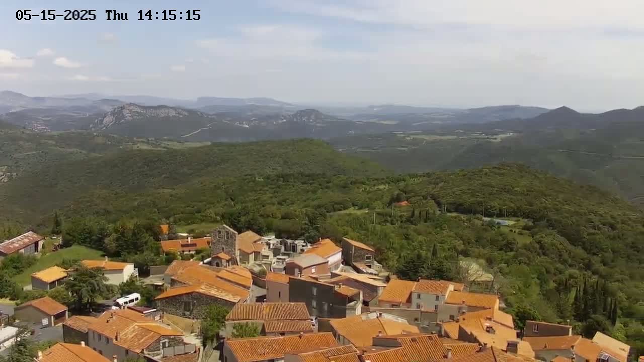 An aerial view shows a small village of orange-roofed buildings nestled in a lush green hillside, overlooking a vast valley and distant mountains under a mostly clear sky.