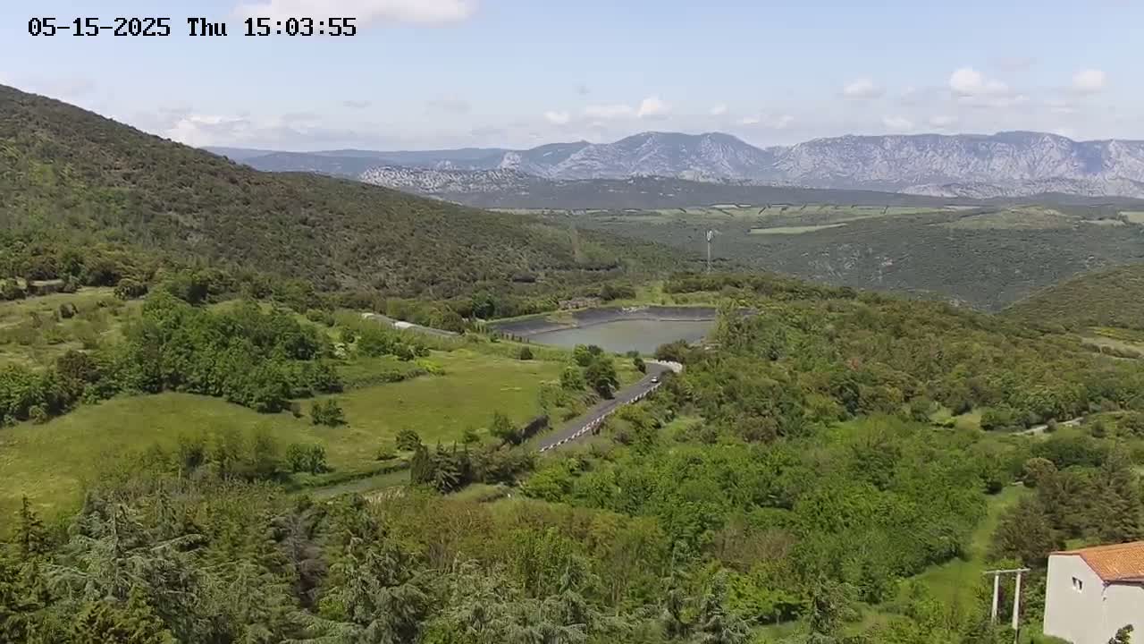 A high-angle view shows a lush green landscape with a small pond, a winding road, and distant mountains under a mostly clear sky.